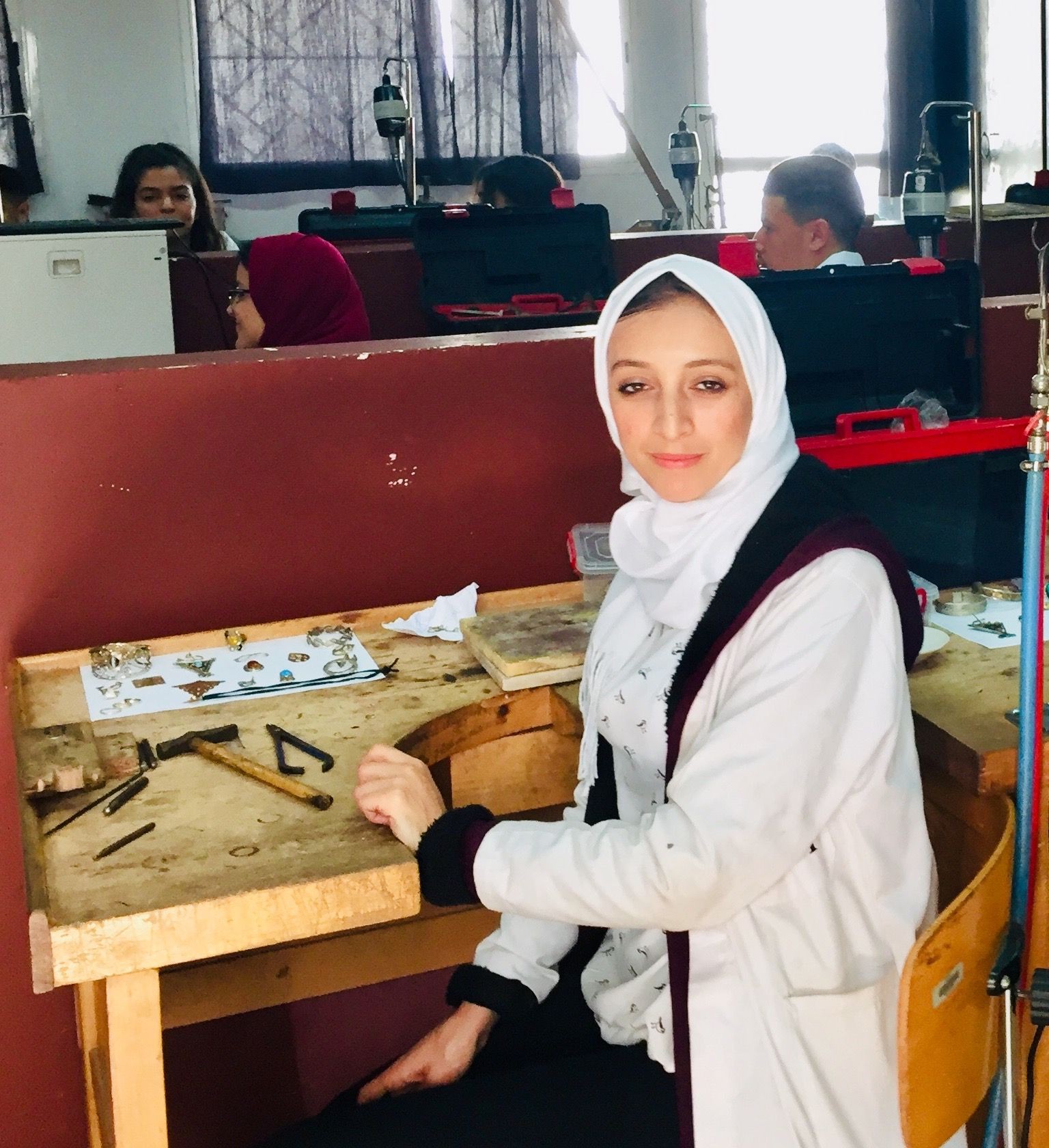 Woman in a white headscarf sitting at a workbench with jewelry tools.